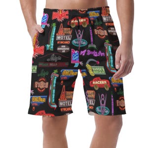 May include: Black shorts patterned with neon signs. The design features signs with text including "MOTEL", "TIRES", and "RACERS". The shorts have a drawstring and pockets.