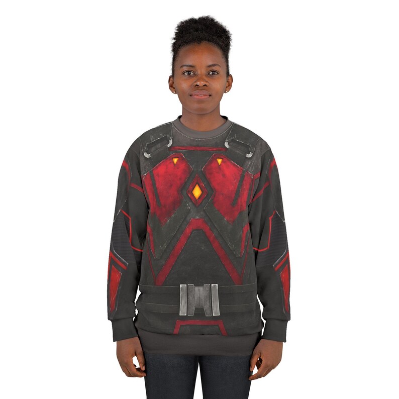 May include: A dark gray sweatshirt with a red and black design resembling armor. The design includes geometric shapes, a belt detail, and a yellow diamond shape. The long-sleeved shirt is worn by a person.