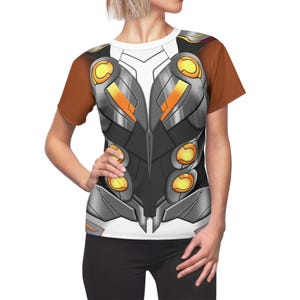 Puede incluir: Una camiseta blanca con un diseño marrón y naranja de una armadura futurista con detalles amarillos brillantes.