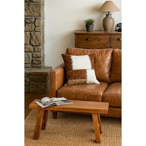 May include: A living room scene with a brown leather sofa and decorative cushions. A small wooden bench holds an open book. A stone fireplace and wooden dresser with a lamp and plant complete the rustic aesthetic.