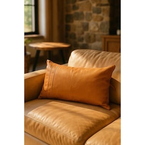 May include: A rectangular, tan leather pillow with vertical stitched detailing on the left side rests on a matching leather sofa. The pillow and sofa are illuminated by natural light, creating a warm, inviting atmosphere. The pillow is approximately 24 inches long.