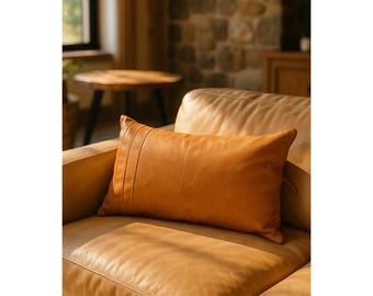 Puede incluir: Un cojín rectangular de cuero marrón claro con detalles de costura verticales en el lado izquierdo descansa sobre un sofá de cuero a juego. El cojín y el sofá están iluminados por luz natural, creando un ambiente cálido. El cojín mide unos 60 cm de largo.