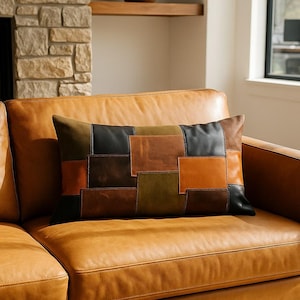 May include: A rectangular cushion with a patchwork design in shades of brown, black, and olive green. The cushion is placed on a tan leather sofa, creating a warm and inviting aesthetic.
