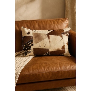 May include: Rectangular decorative pillow with a patchwork design. The pillow features various shades of brown and cream-colored leather pieces, creating a geometric pattern. It rests on a brown leather armchair, with a cream-colored knitted throw visible.