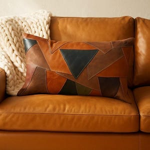May include: A rectangular cushion with a patchwork design in shades of brown, tan, and black. The cushion is placed on a brown leather sofa, with a cream-coloured knitted throw blanket visible in the background.