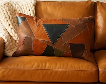 May include: Rectangular decorative pillow with a patchwork design in shades of brown, tan, and black. The pillow is placed on a brown leather sofa, with a cream-colored knitted throw blanket visible in the background.