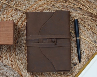 May include: A brown leather journal with a wrap-around closure. A black pen rests beside the journal on a woven mat. The journal's cover has a unique design, and the closure is secured with a leather strap.