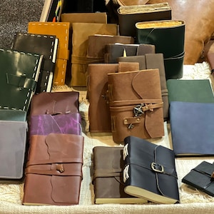 Pode incluir: Uma coleção de diários encadernados em couro com várias cores e designs. Alguns diários têm correias e fivelas decorativas.