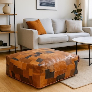 Op de afbeelding: Vierkante leren poef met een patchwork ontwerp in bruin- en donkergrijstinten. De poef staat op een houten vloer in een woonkamer. Een lichtgrijze bank en een ronde salontafel staan op de achtergrond.