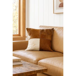 May include: Square decorative pillow with a patchwork design. The pillow features a combination of brown and cream-colored leather squares, stitched together with visible seams. The pillow is placed on a tan leather sofa.