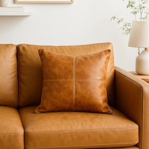 May include: A square, tan leather pillow with stitched detailing, placed on a matching leather sofa. The pillow has a cross-stitched design. The sofa is part of a living room set.