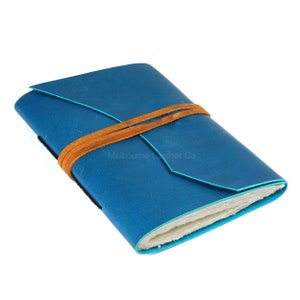 Könnte beinhalten: Ein blaues Lederjournal mit einem braunen Lederriemenverschluss. Das Journal ist geschlossen und der Riemen ist zu einem Knoten gebunden. Der Text "Melbourne Leather Co." ist auf dem Einband aufgedruckt.