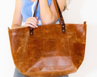 May include: A large, brown leather tote bag with a rectangular shape and a front pocket. The bag has two dark brown leather handles attached with metal rivets. The leather has a distressed, textured appearance.