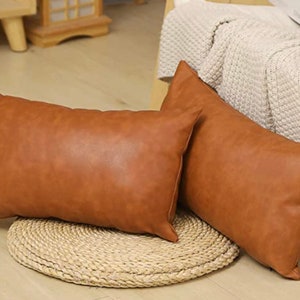 May include: Two brown leather-look throw pillows with a rectangular shape. The pillows are on a woven rug.