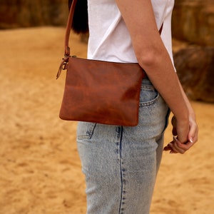 May include: A brown leather crossbody bag with a zipper closure. The bag is worn over the shoulder of a person wearing a white t-shirt and blue jeans.