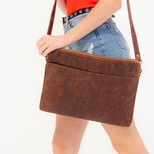 May include: A brown leather laptop bag with a flap closure and a front pocket. The bag is worn over the shoulder of a person wearing a red shirt and denim shorts.