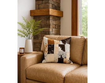 May include: A tan leather sofa with a decorative patchwork pillow. The pillow features squares of brown, black, and cream-colored cowhide. A white vase with green fern and a small candle sit on a side table.