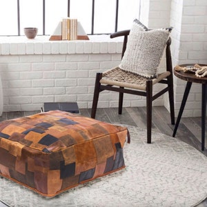 Puede incluir: Un gran otomano cuadrado de cuero patchwork en tonos marrones, negros y beige. El otomano está sobre una alfombra gris claro en una habitación con paredes de ladrillo blanco.