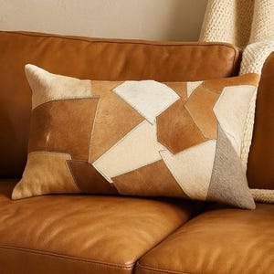 Könnte beinhalten: Rechteckiges Zierkissen mit Patchwork-Design. Das Kissen hat verschiedene Brauntöne, Beige und Creme, die aus Leder- oder Fellstücken zusammengenäht sind. Es liegt auf einem braunen Ledersofa, mit einer neutralfarbenen Decke im Hintergrund.