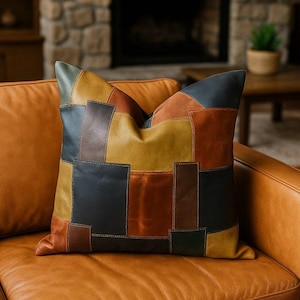 Peut inclure: Un coussin carré décoratif avec un motif patchwork. Il présente des pièces de cuir rectangulaires et carrées dans des tons de marron, noir, vert et jaune. Le coussin est posé sur un canapé en cuir marron.