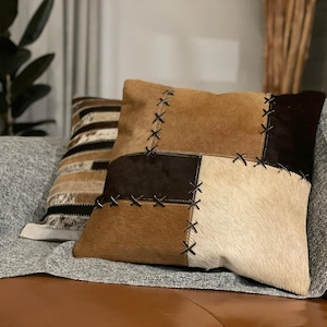 May include: A brown, white and black patchwork cushion with decorative stitching. The cushion is made of different types of leather and has a soft, luxurious feel.