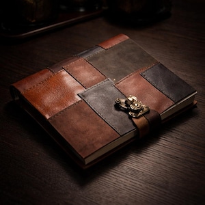 May include: A leather journal with a patchwork design in shades of brown and gray. The journal features a clasp closure and a leather strap. The cover is made of rectangular pieces of leather stitched together. The journal is a dark brown color.