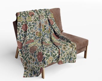 May include: A floral patterned throw blanket draped over a brown chair. The blanket features a repeating design of roses, leaves, and birds in shades of red, green, blue, and tan. The chair has a wooden frame.