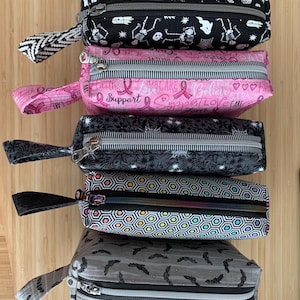 Peut inclure: Une pile de cinq trousses rectangulaires avec fermetures éclair et dragonnes. Les trousses présentent divers motifs, notamment des squelettes, des rubans roses, des formes géométriques et des chauves-souris. Chaque trousse a une palette de couleurs différente, comme le noir, le rose, le gris et multicolore.