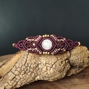May include: A handmade macrame bracelet featuring a central white gemstone. The bracelet is crafted with deep purple and tan cords, accented with small gold-colored beads. The bracelet is displayed on a piece of textured wood.