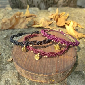 Könnte beinhalten: Drei geflochtene Armbänder mit goldenen Anhängern. Die Armbänder sind schwarz, burgunderrot und eine Kombination aus beiden Farben. Die Anhänger sind rund und quadratisch.
