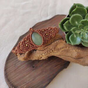 Peut inclure: Un bracelet en macramé marron avec une pierre précieuse verte et des perles dorées.