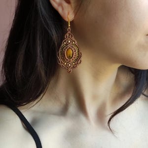 Può includere: Un orecchino in macramè fatto a mano a forma di goccia. L'orecchino presenta una pietra centrale ovale color ambra, circondata da intricati dettagli in macramè marrone e oro. L'orecchino è appeso a un gancio color oro.