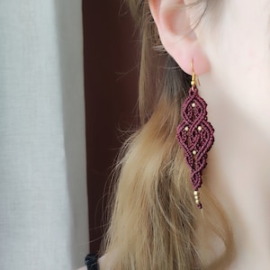 May include: Burgundy red macrame earrings with gold beads. The earrings are in the shape of a teardrop and have a delicate, intricate design.