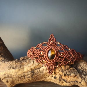 Könnte beinhalten: Ein braunes Makramee-Diadem mit einem ovalen Tigeraugenstein in der Mitte. Das Diadem ist mit kleinen goldenen Perlen und aufwendigen Knotenarbeiten verziert. Es wird auf einem Stück Treibholz präsentiert.