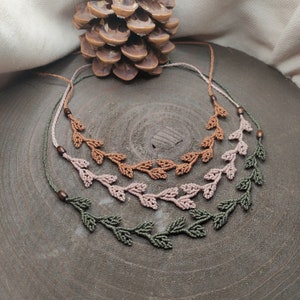 May include: Three macrame necklaces with leaf designs in shades of green, pink and brown. The necklaces are displayed on a wooden surface.