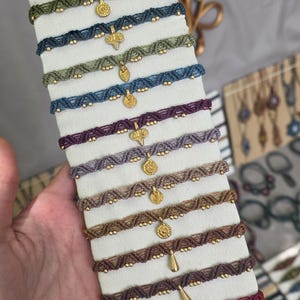 May include: A selection of handmade macrame chokers in shades of green, blue, purple, and brown. Each choker is adorned with gold-coloured beads and a gold pendant. The chokers are displayed on a white stand.