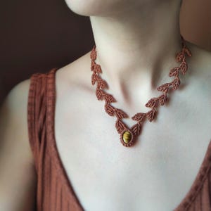 May include: A brown macrame necklace featuring a leaf design. The necklace has a central oval-shaped pendant with a yellow-brown stone. The leaves are intricately knotted, creating a delicate and natural aesthetic. The necklace is worn around the neck.