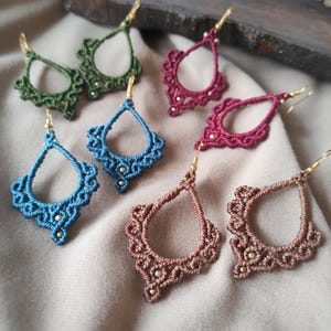 Può includere: Un set di sei orecchini all'uncinetto fatti a mano in vari colori, tra cui turchese, bordeaux, marrone e verde. Ogni orecchino presenta una forma a goccia con dettagli intricati e un gancio dorato.