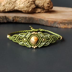 Puede incluir: Pulsera de macramé hecha a mano con una piedra central redonda de color marrón claro. La pulsera está elaborada con cordón verde oliva y verde claro, adornada con pequeñas cuentas doradas. El diseño es intrincado y detallado, de estilo bohemio.