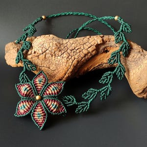 May include: A green macrame necklace with a flower pendant. The flower is made of green, red, and gold cord and has a gold bead in the center.