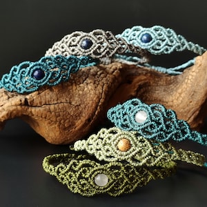 May include: Several macrame bracelets in shades of teal, olive green, and grey. Each bracelet has a central gemstone, like a blue lapis lazuli or a white quartz. The bracelets are presented on a piece of driftwood, against a dark background.