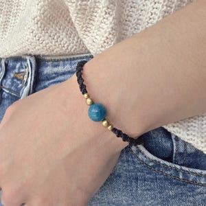 May include: Black macrame bracelet featuring a large, round, blue gemstone bead and small gold-colored beads. The bracelet is worn on a wrist, with a light-colored sweater and blue jeans in the background. A simple, elegant piece of jewelry.