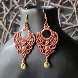 Könnte beinhalten: Ein Paar handgefertigte Makramee-Ohrringe in einem rötlich-braunen Farbton. Die Ohrringe weisen komplizierte Knotenmuster, kleine goldfarbene Perlen und ein kreisförmiges Design auf. Die Ohrringe haben goldfarbene Ohrhaken und einen dekorativen goldfarbenen Anhänger.