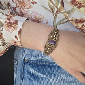 Può includere: Un braccialetto in macramè con una pietra centrale di ametista ovale e piccole perline color oro. Il braccialetto è di colore marrone caldo e viene indossato su un polso. Lo sfondo include una camicetta a fantasia floreale e jeans.