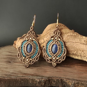 Puede incluir: Un par de pendientes de macramé hechos a mano. Los pendientes tienen forma de lágrima con una piedra azul ovalada en el centro, rodeada de macramé turquesa y marrón. Cuentas doradas y ganchos dorados completan el diseño.