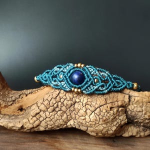 May include: A teal macrame bracelet with a central dark blue gemstone and gold-coloured beads. The bracelet is displayed on a piece of textured wood against a dark background. The macrame design features intricate knotting.