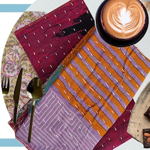 May include: An assortment of colorful, patterned fabric napkins with white stitching, alongside gold cutlery and a cup of coffee with latte art. The napkins feature stripes, floral prints, and geometric designs, creating a vibrant table setting.