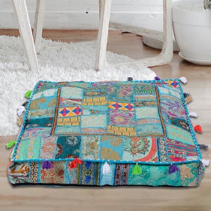 Puede incluir: Un cojín de suelo azul turquesa con un diseño de patchwork hecho de diferentes telas. El cojín tiene un borde de flecos y está decorado con bordados coloridos.