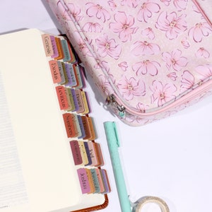 May include: A pink floral zippered case with a white zipper and a white and brown bible with colorful tabs labeled with book names from the Old Testament, including Genesis, Exodus, Leviticus, Numbers, Deuteronomy, Joshua, Judges, Ruth, 1 Samuel, 2 Samuel, 1 Kings, 2 Kings, 1 Chronicles, 2 Chronicles, Ezra, Nehemiah, Esther, Job, Psalms, Proverbs, Ecclesiastes, Song of Solomon, Isaiah, Jeremiah, Lamentations, Ezekiel, Daniel, Hosea, Joel, Amos, Obadiah, Jonah, Micah, Nahum, Habakkuk, Zephaniah, Haggai, Zechariah, Malachi.