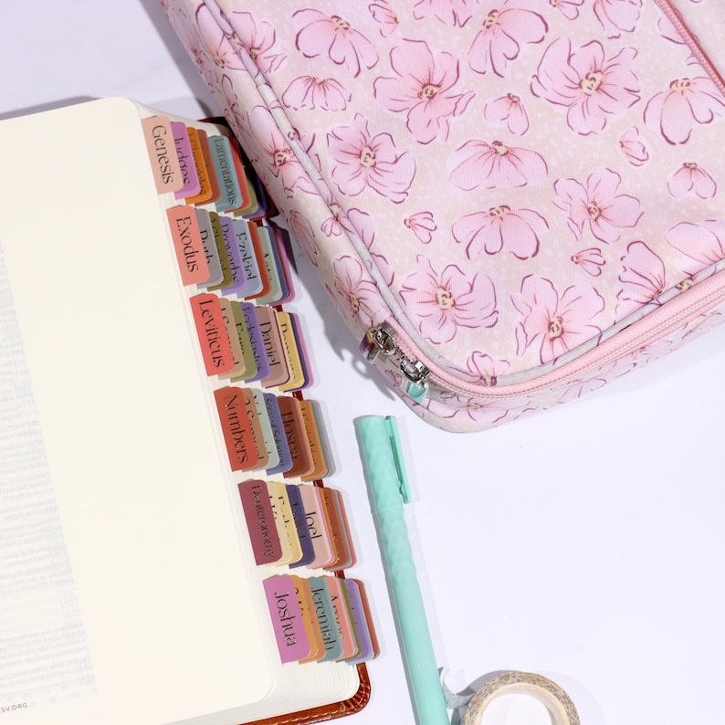 May include: A pink floral zippered case with a white zipper and a white and brown bible with colorful tabs labeled with book names from the Old Testament, including Genesis, Exodus, Leviticus, Numbers, Deuteronomy, Joshua, Judges, Ruth, 1 Samuel, 2 Samuel, 1 Kings, 2 Kings, 1 Chronicles, 2 Chronicles, Ezra, Nehemiah, Esther, Job, Psalms, Proverbs, Ecclesiastes, Song of Solomon, Isaiah, Jeremiah, Lamentations, Ezekiel, Daniel, Hosea, Joel, Amos, Obadiah, Jonah, Micah, Nahum, Habakkuk, Zephaniah, Haggai, Zechariah, Malachi.